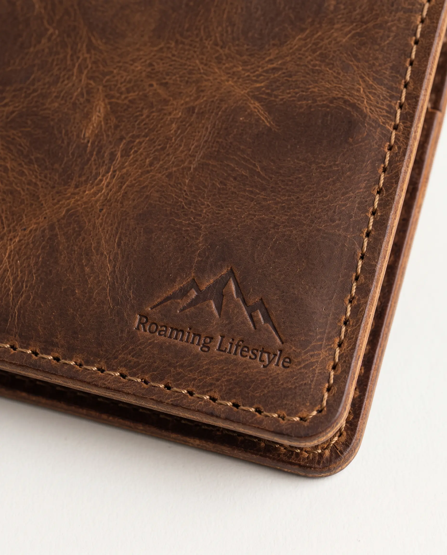 Close-up detail of tobacco brown full-grain leather wallet showing debossed mountain crest logo, hand-burnished edge, and contrast perimeter stitching – Roaming Lifestyle