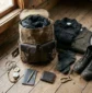 Flat lay of a packed waxed canvas rucksack, wool tweed vest, rugged leather boots, notebook, and brass pocket compass on a rustic wooden floor – Roaming Lifestyle