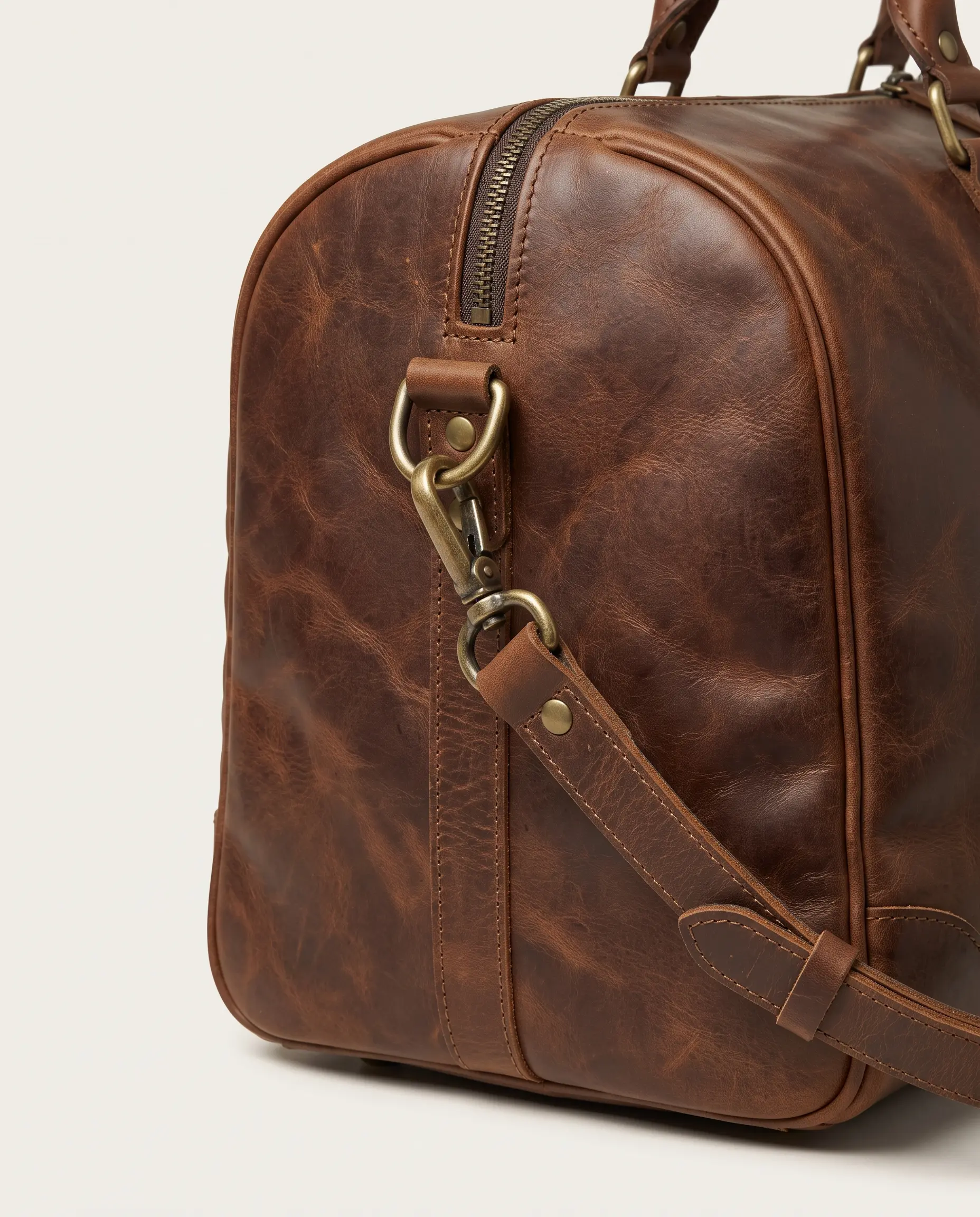 Close-up side view of brown leather weekender duffle bag showing antique brass lobster clasp, D-ring, rivets, zippered edge, and textured stitching on plain studio background – Roaming Lifestyle
